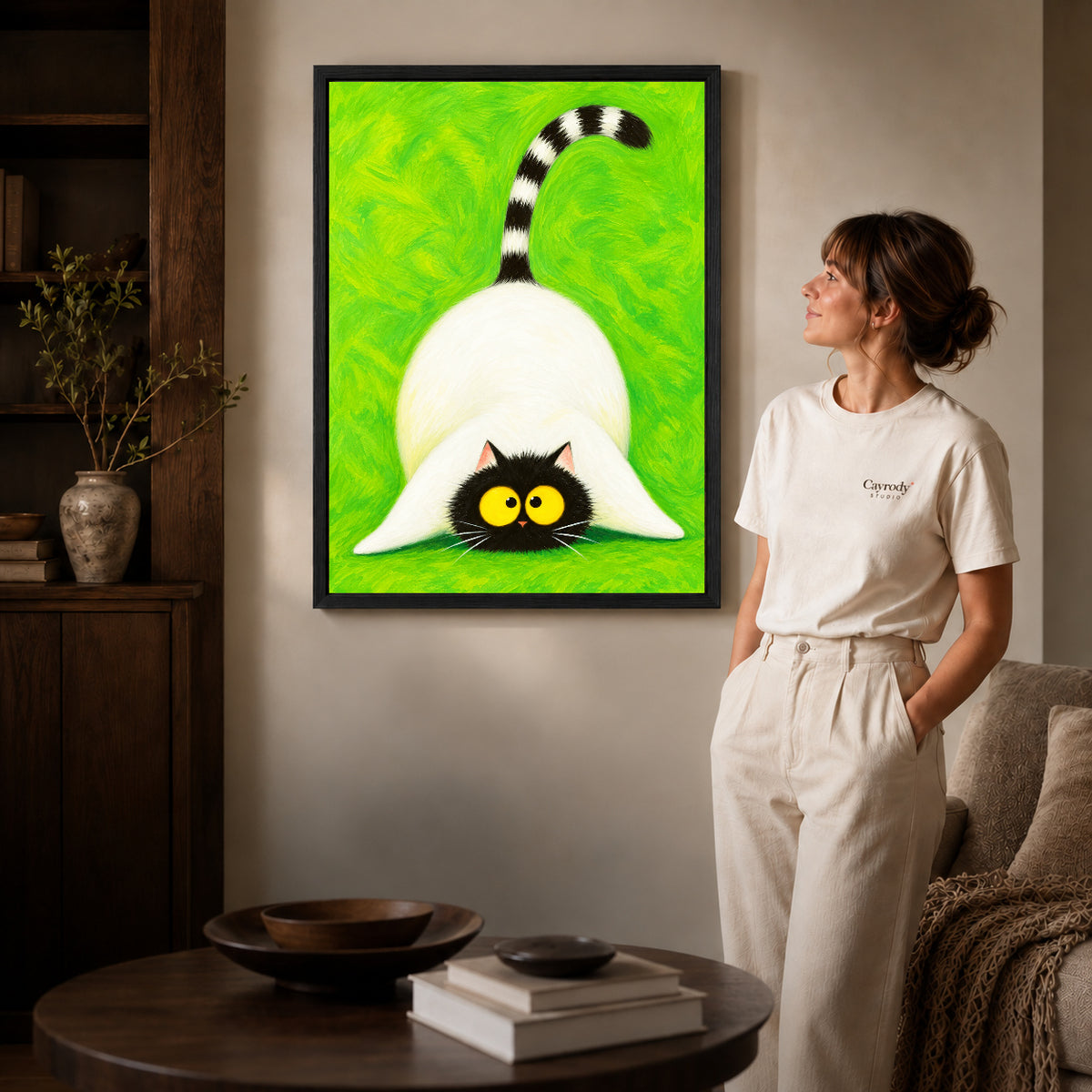 Playful cat canvas art in a black frame beside a woman and round wooden table.