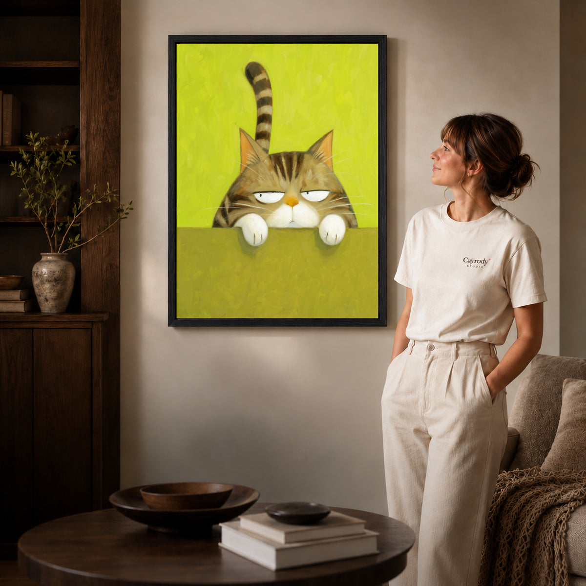 Grumpy tabby canvas art in a black frame beside a woman and round wooden table.