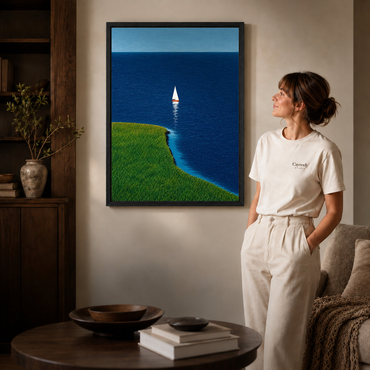 Coastal sailboat canvas art in a black frame beside a woman and round wooden table.