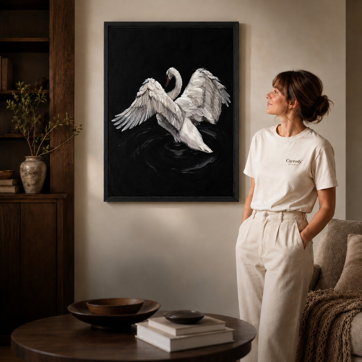 White swan canvas art in a black frame beside a woman and round wooden table.