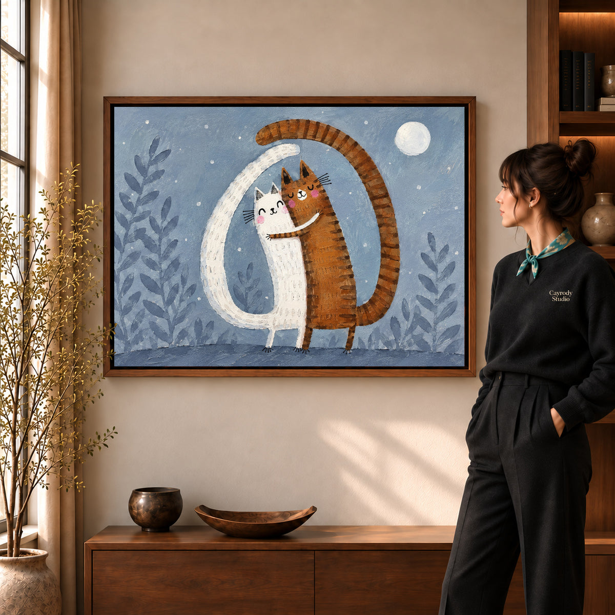 Hugging cats canvas art in a walnut frame beside a standing woman and wooden console.