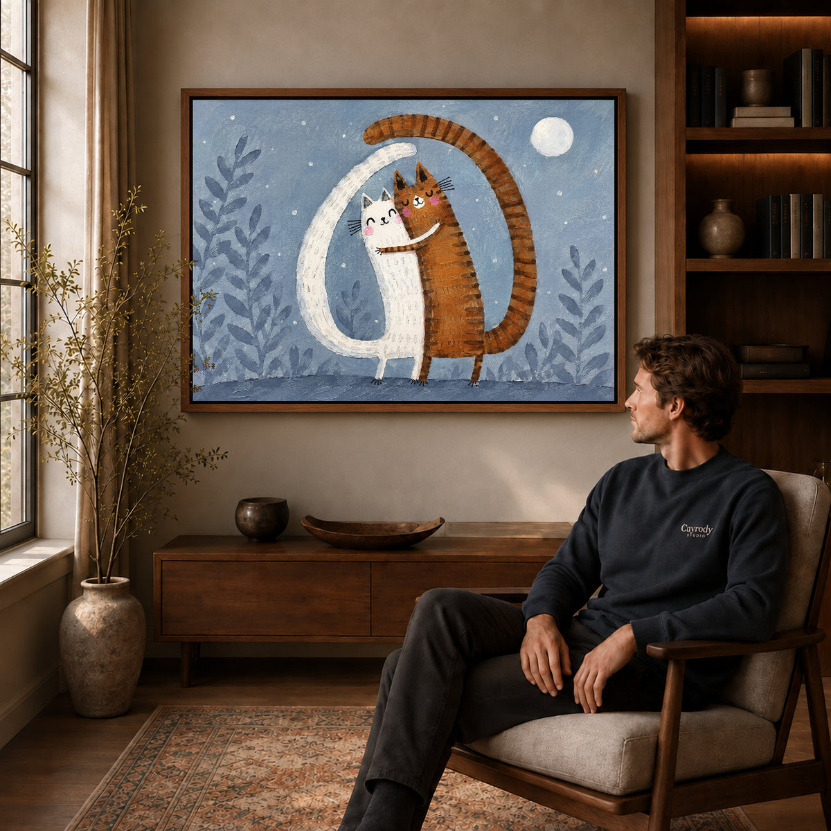 Hugging cats canvas art in a walnut frame above a console beside a seated man.