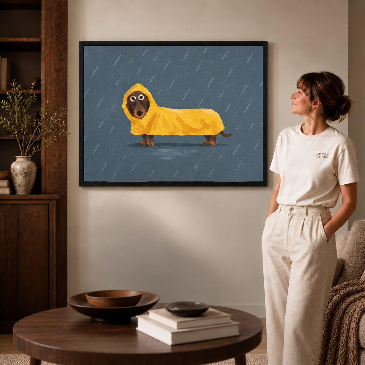 Dachshund raincoat canvas art in a black frame beside a woman and round wooden table.
