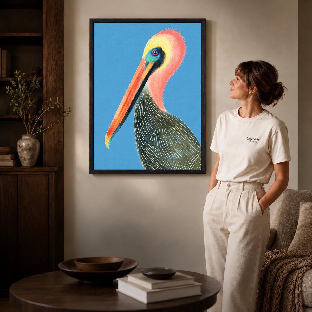Colorful pelican canvas art in a black frame beside a woman and round wooden table.