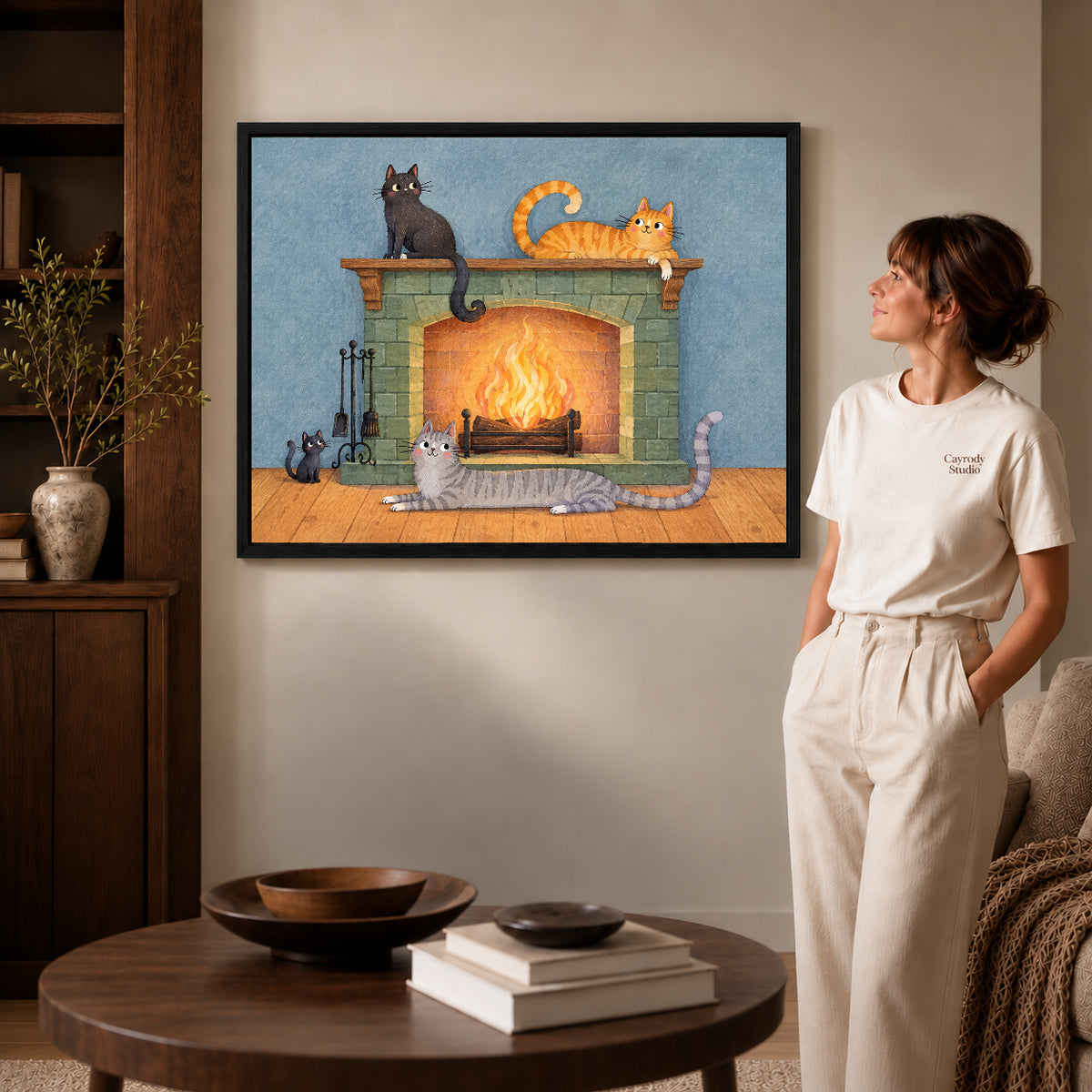 Fireplace cats canvas art in a black frame beside a woman and round wooden table.