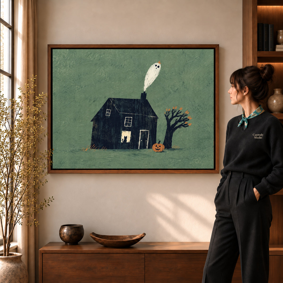 Ghost house canvas art in a walnut frame beside a standing woman and wooden console.
