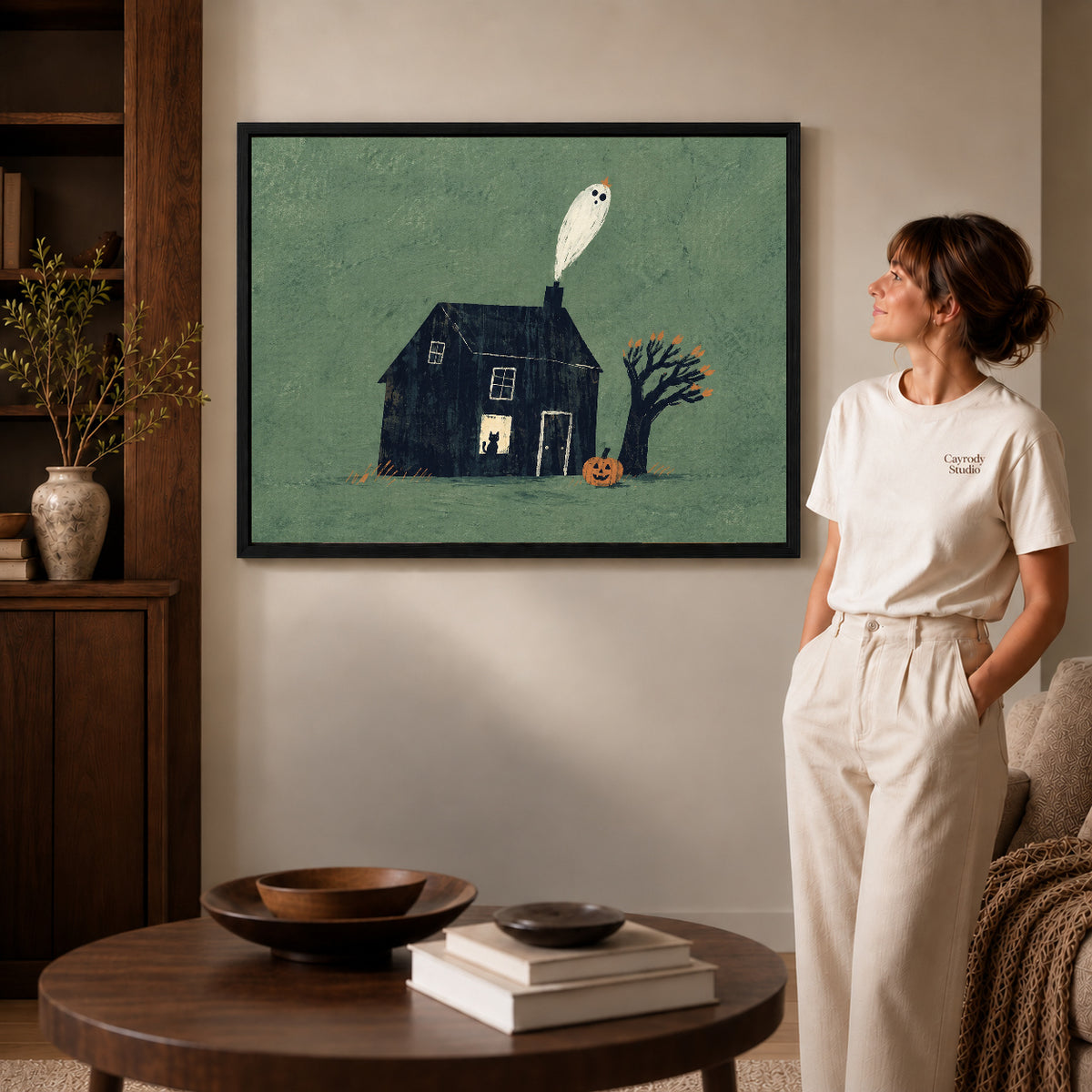 Ghost house canvas art in a black frame beside a woman and round wooden table.