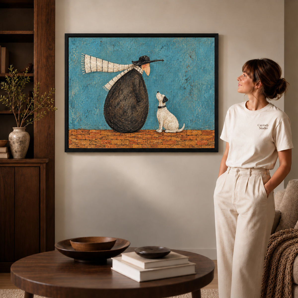 Whimsical companion canvas art in a black frame beside a woman and round wooden table.