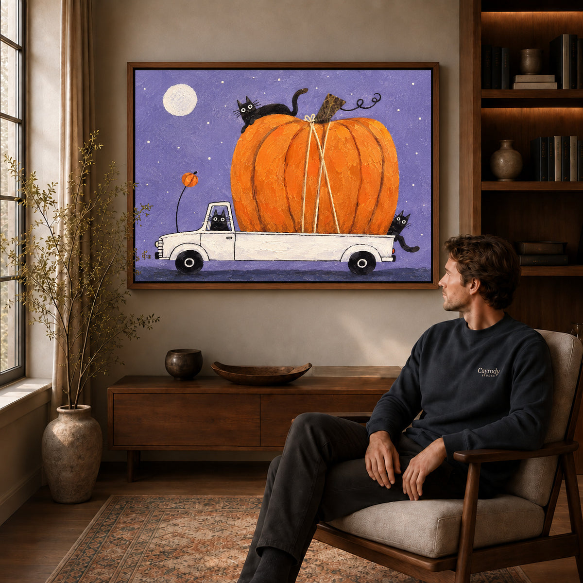Pumpkin truck cats canvas art in a walnut frame above a console beside a seated man.