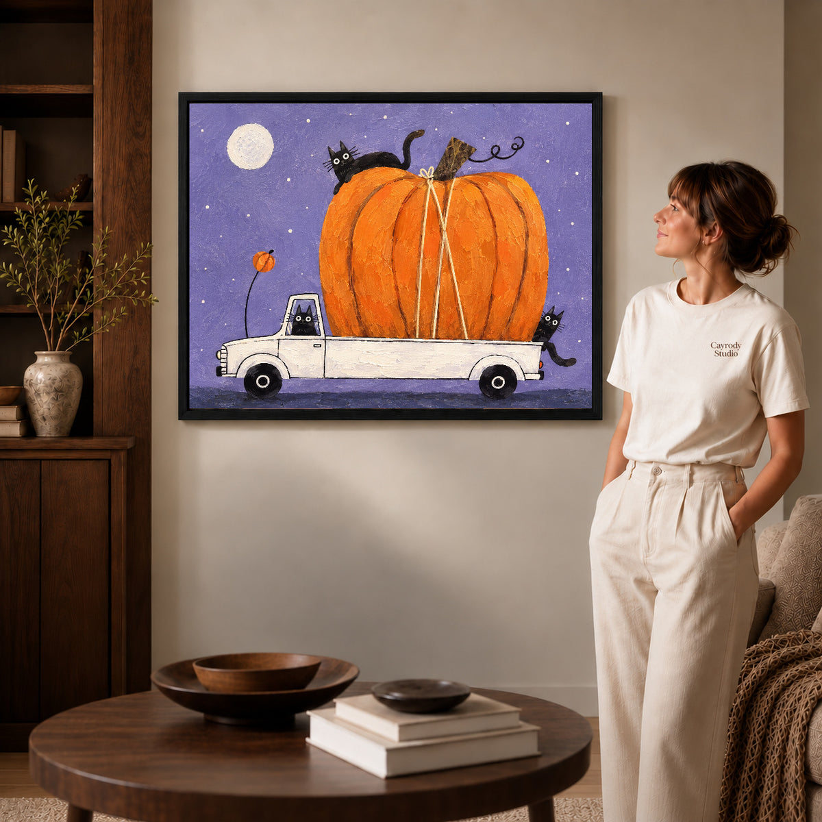 Pumpkin truck cats canvas art shown in the black frame option with warm furnishings and a standing woman.