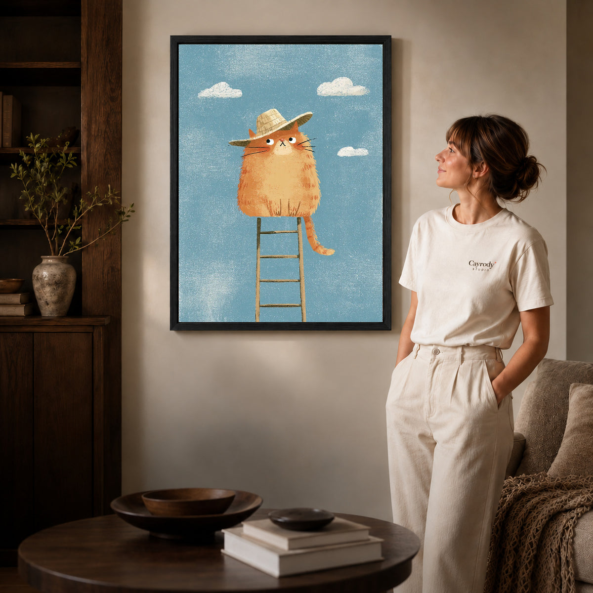 straw hat cat canvas art in a black frame beside a woman and round wooden table.