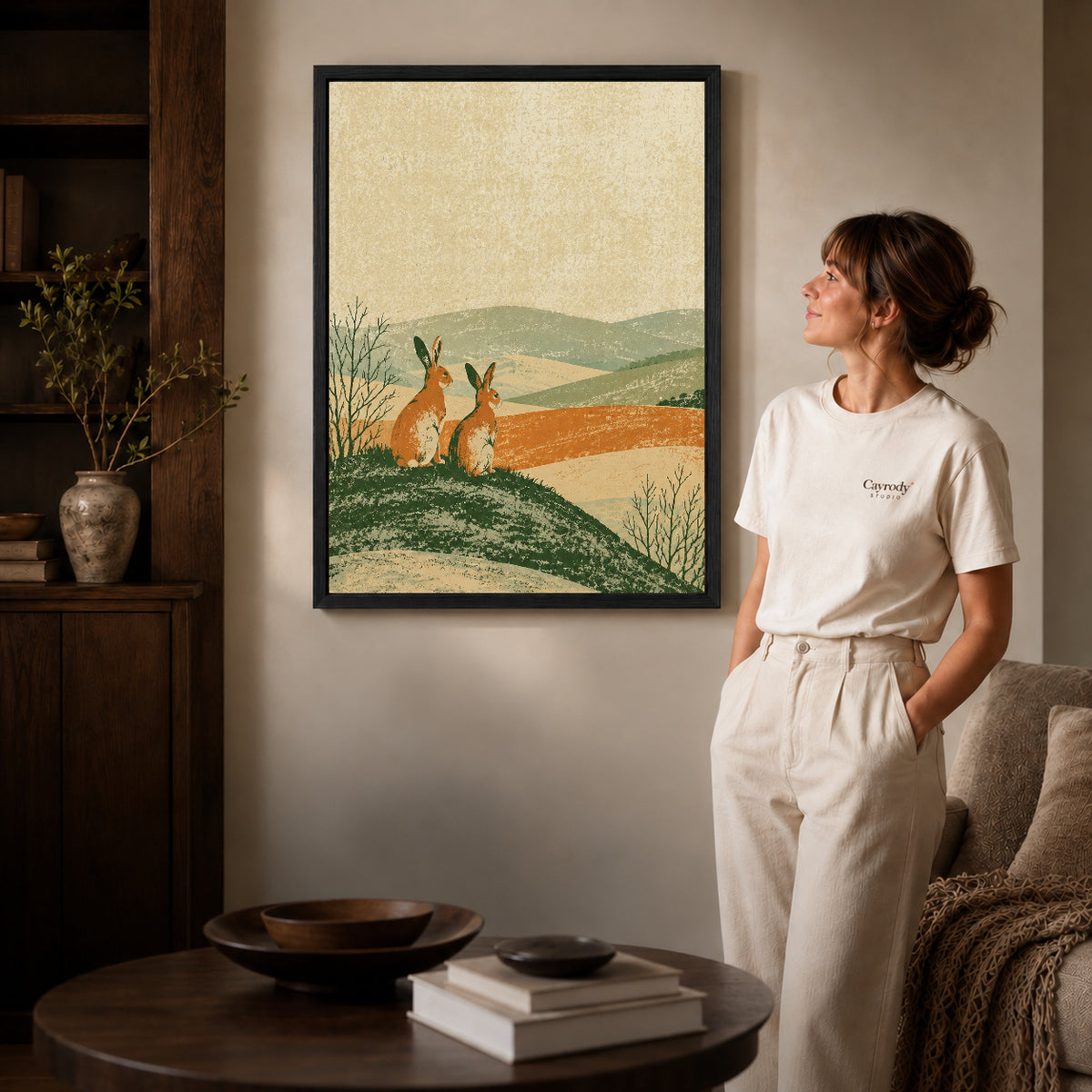 rustic rabbit landscape canvas art in a black frame beside a woman and round wooden table.