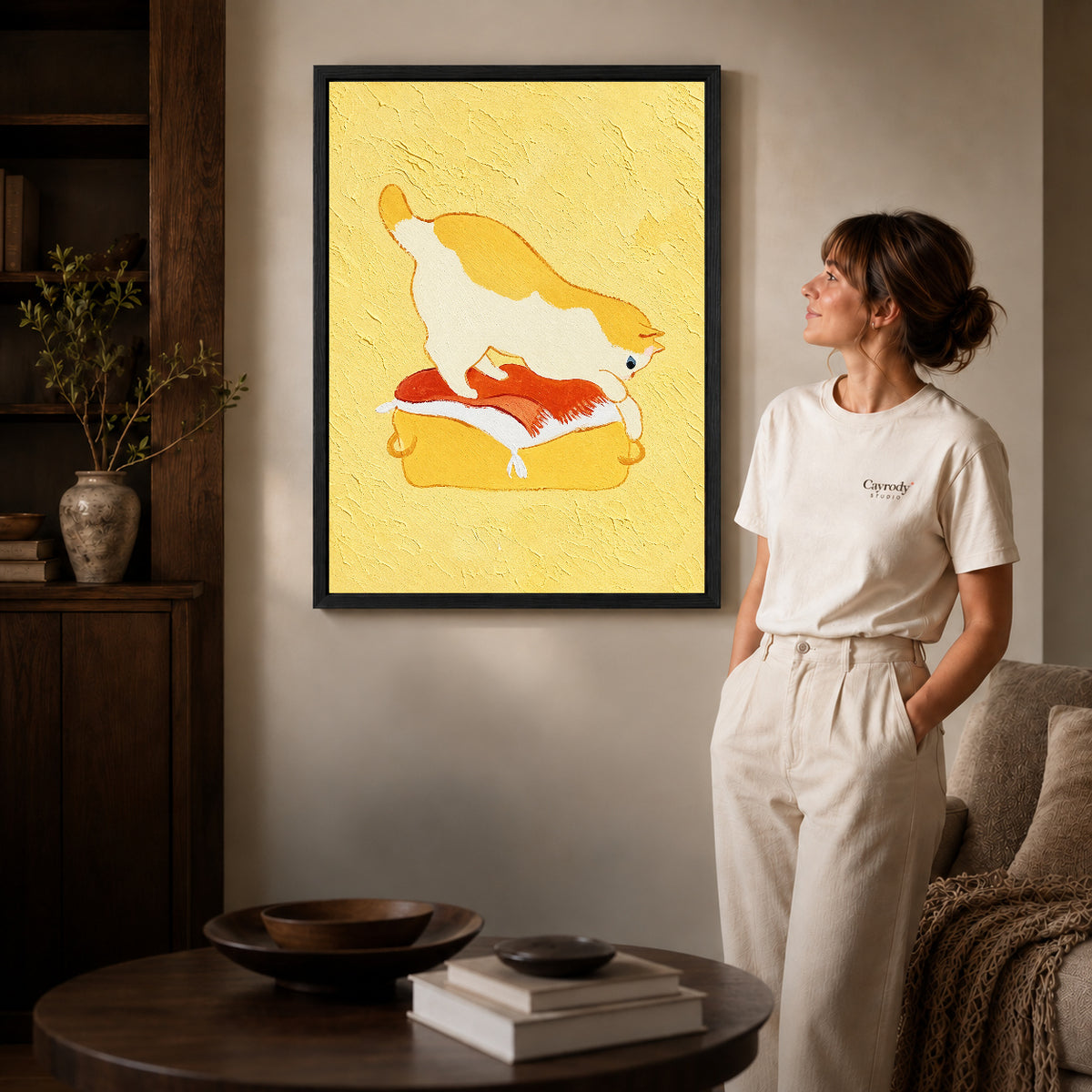 playful kneading cat canvas art in a black frame beside a woman and round wooden table.