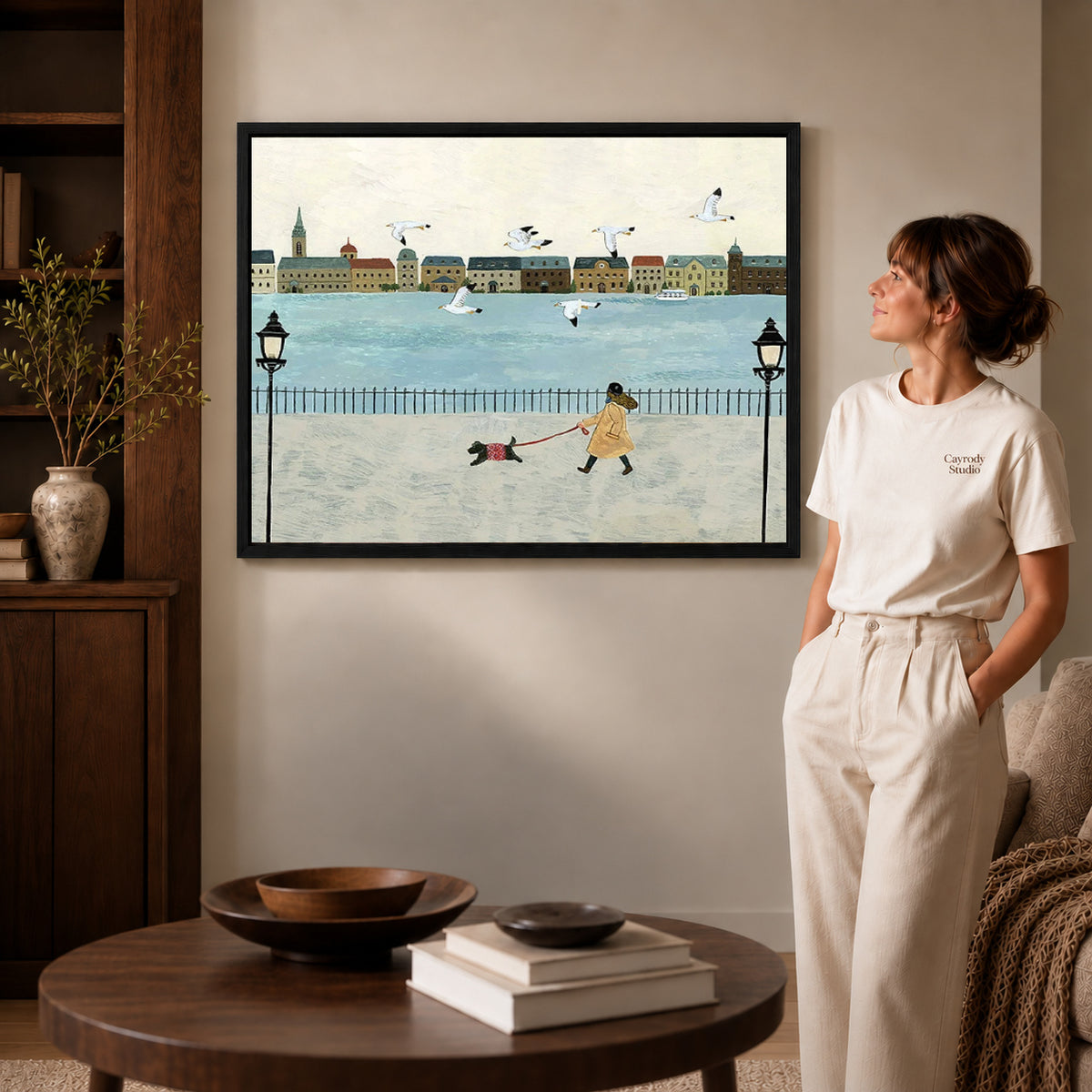folk waterfront walk canvas art in a black frame beside a woman and round wooden table.