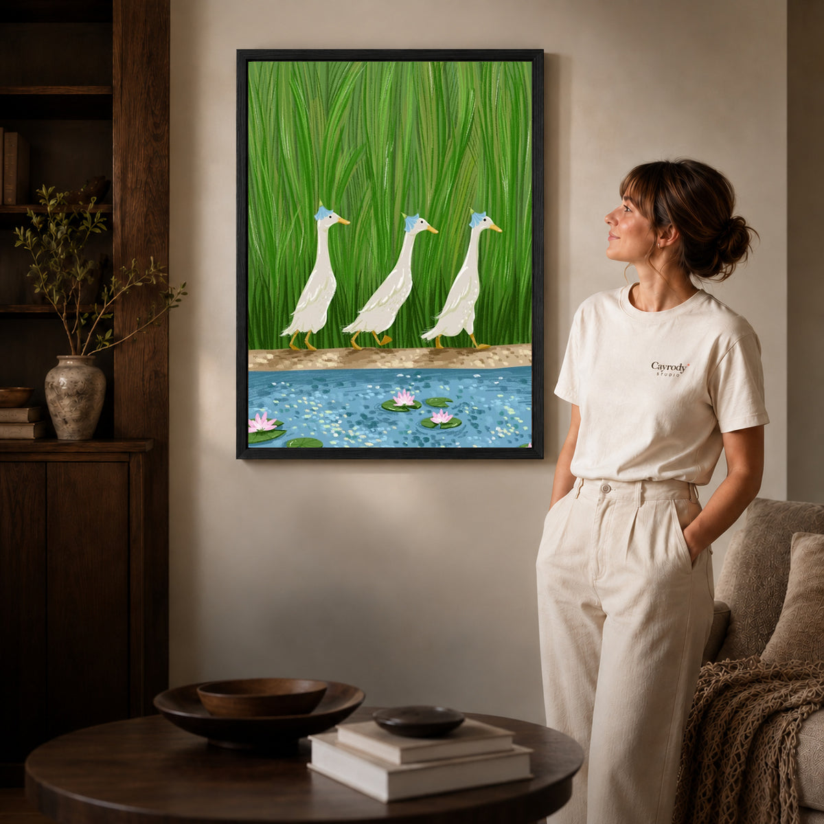 folk pond geese canvas art in a black frame beside a woman and round wooden table.