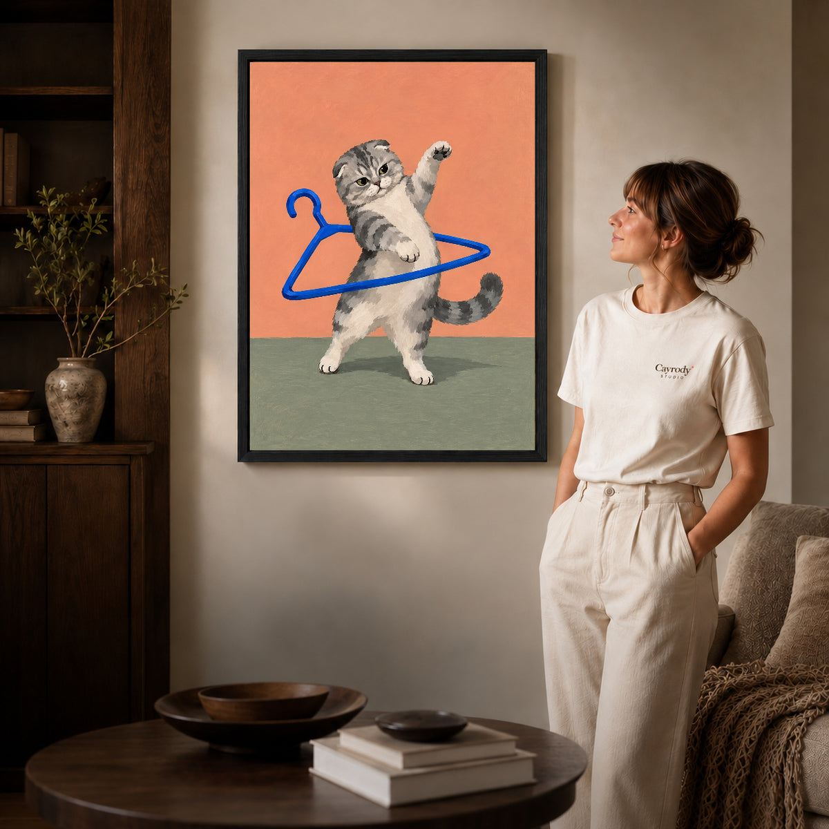playful hanger kitten canvas art in a black frame beside a woman and round wooden table.