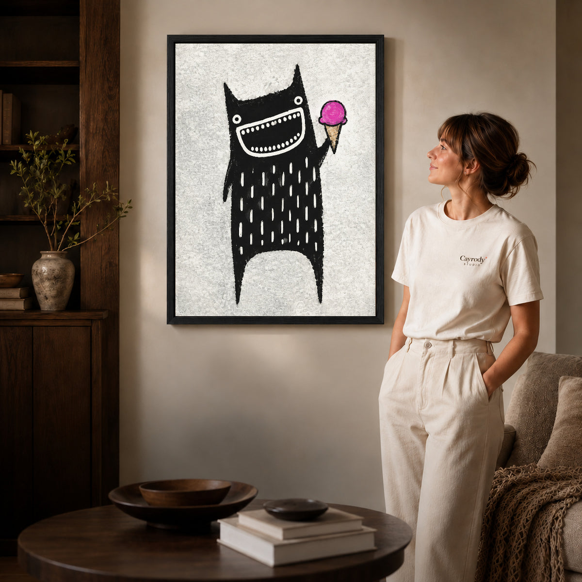 folk ice cream monster canvas art in a black frame beside a woman and round wooden table.