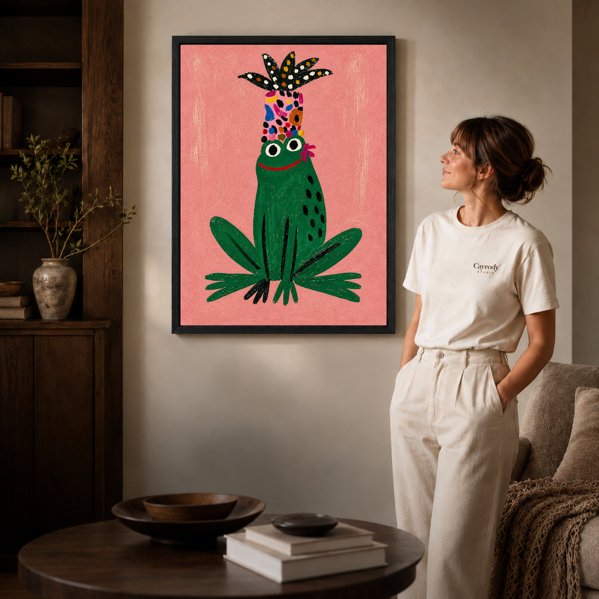 folk botanical frog canvas art in a black frame beside a woman and round wooden table.