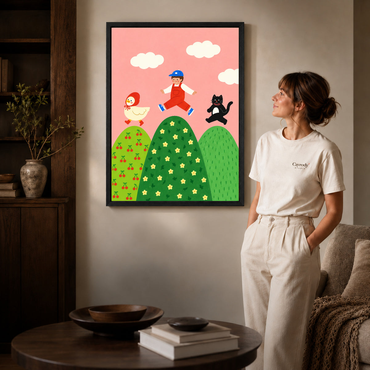 playful hilltop friends canvas art in a black frame beside a woman and round wooden table.