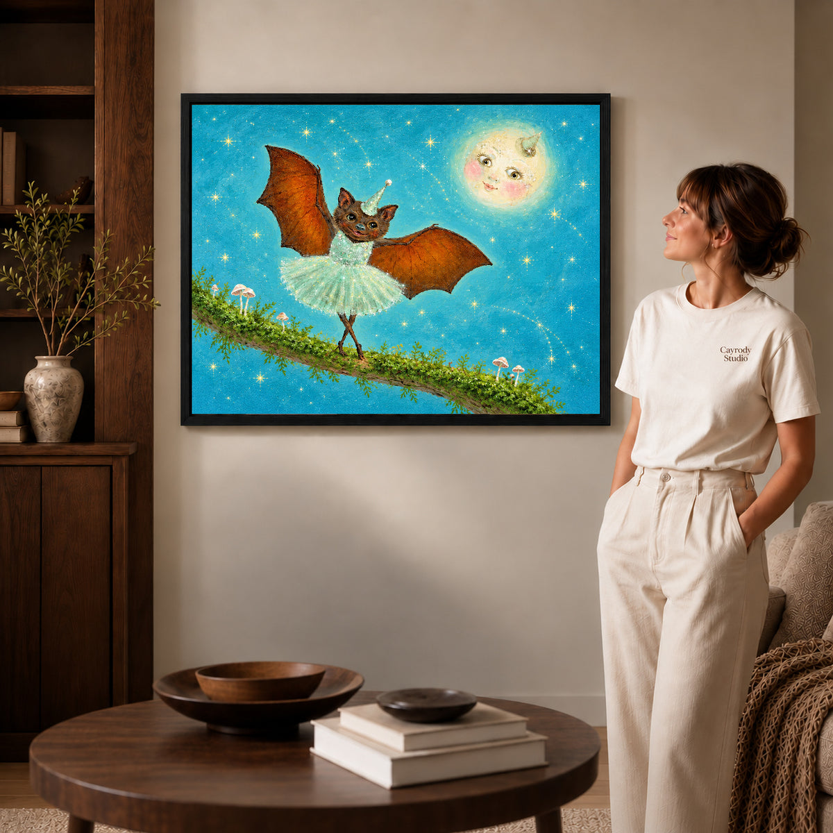 fantasy ballet bat canvas art in a black frame beside a woman and round wooden table.