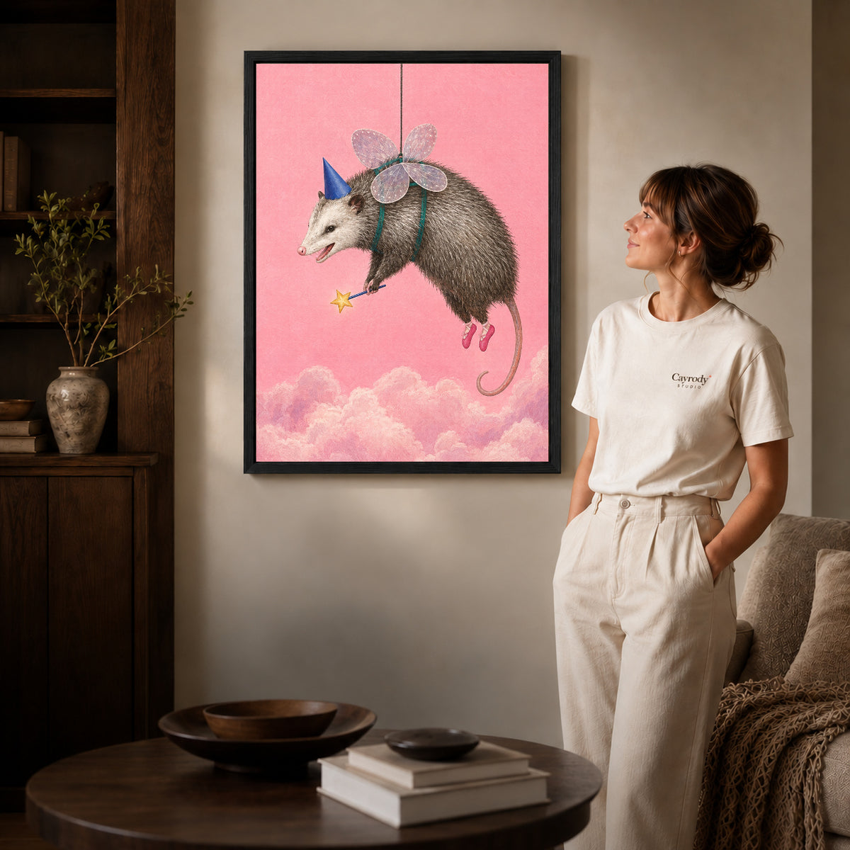 whimsical fairy opossum canvas art in a black frame beside a woman and round wooden table.
