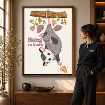 whimsical hanging opossum canvas art in a walnut frame beside a standing woman and wooden console.