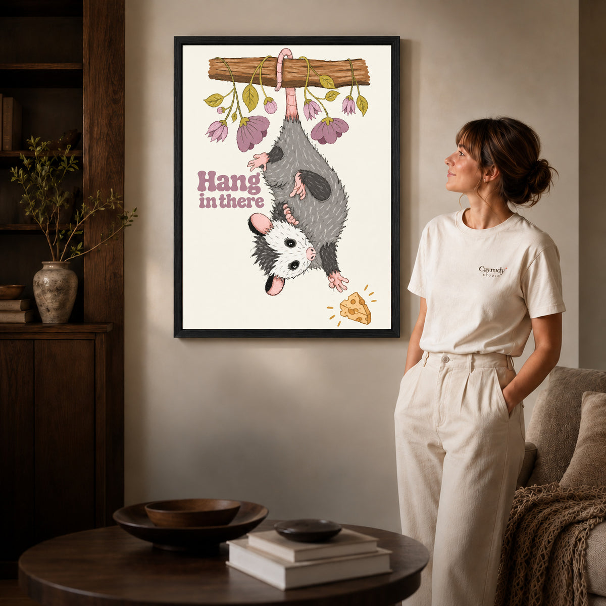 whimsical hanging opossum canvas art in a black frame beside a woman and round wooden table.