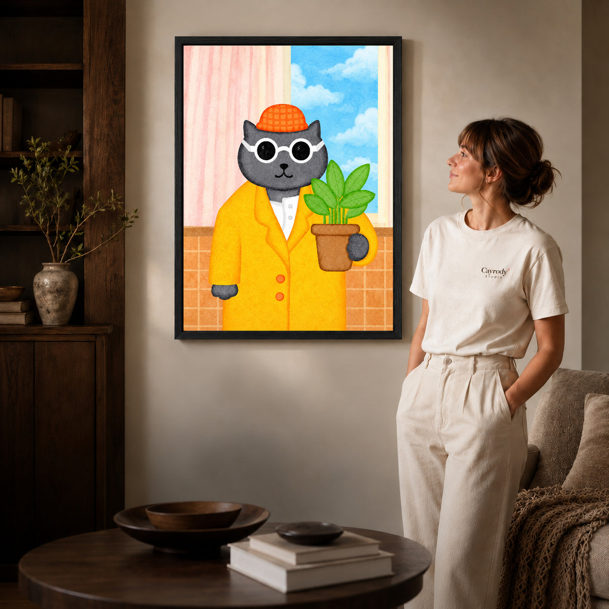whimsical plant lover cat canvas art in a black frame beside a woman and round wooden table.