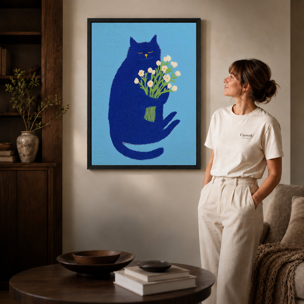 blue cat with flowers canvas art in a black frame beside a woman and round wooden table.