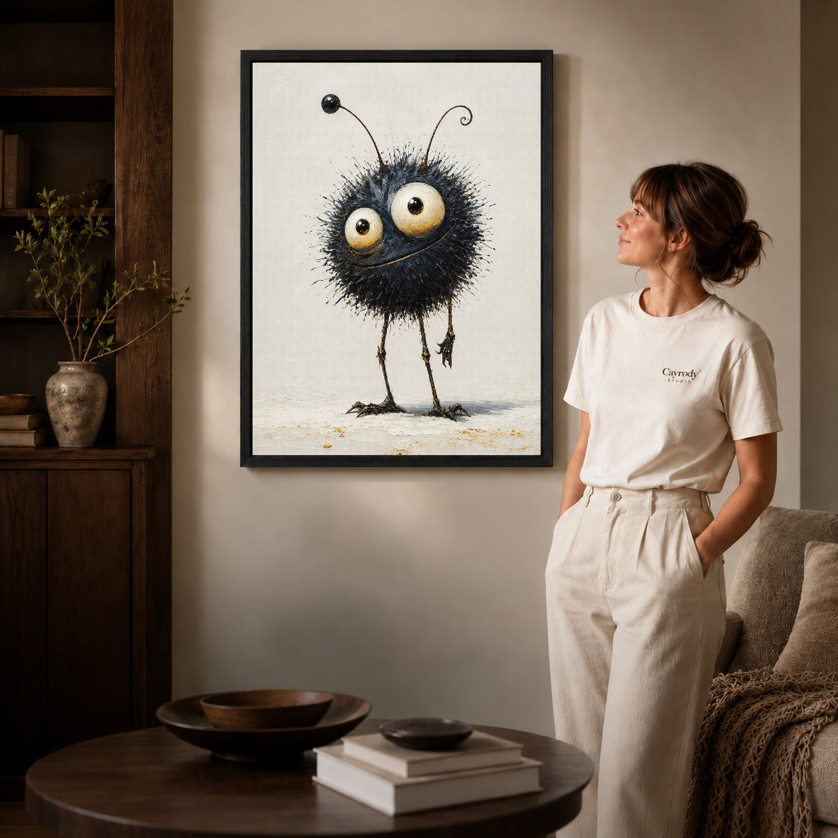 whimsical fuzzy creature canvas art in a black frame beside a woman and round wooden table.