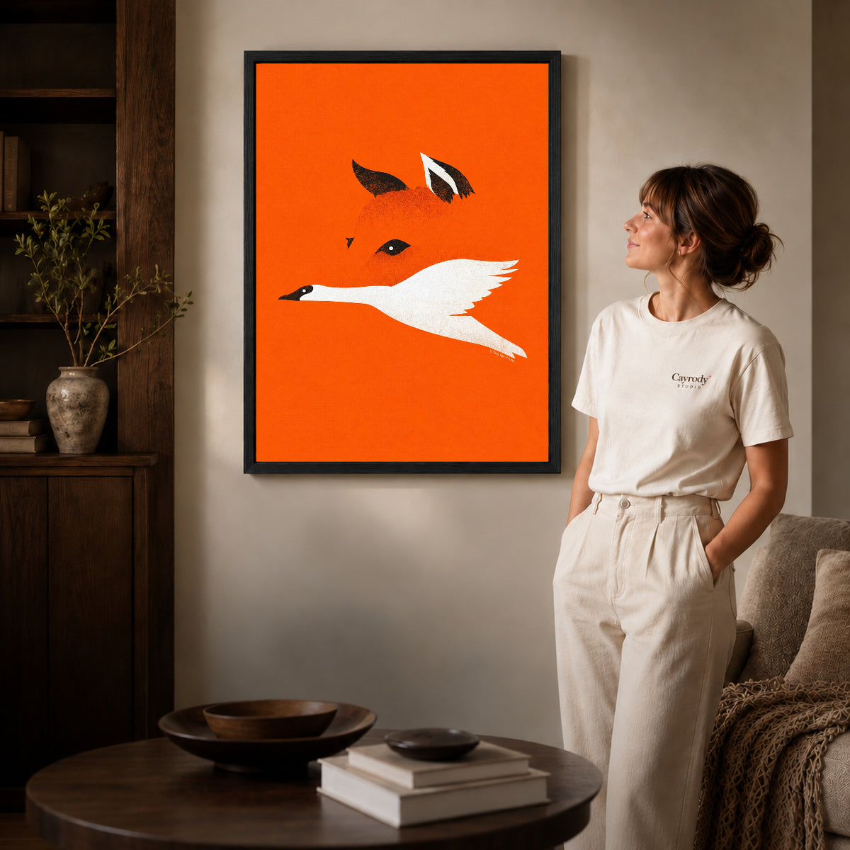 minimalist fox bird canvas art in a black frame beside a woman and round wooden table.
