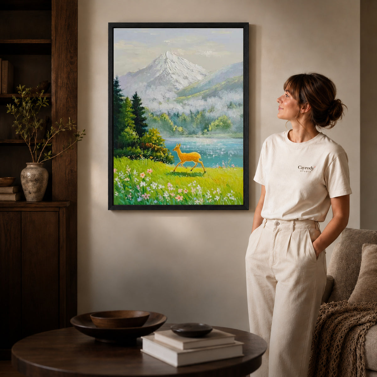 alpine deer meadow canvas art in a black frame beside a woman and round wooden table.