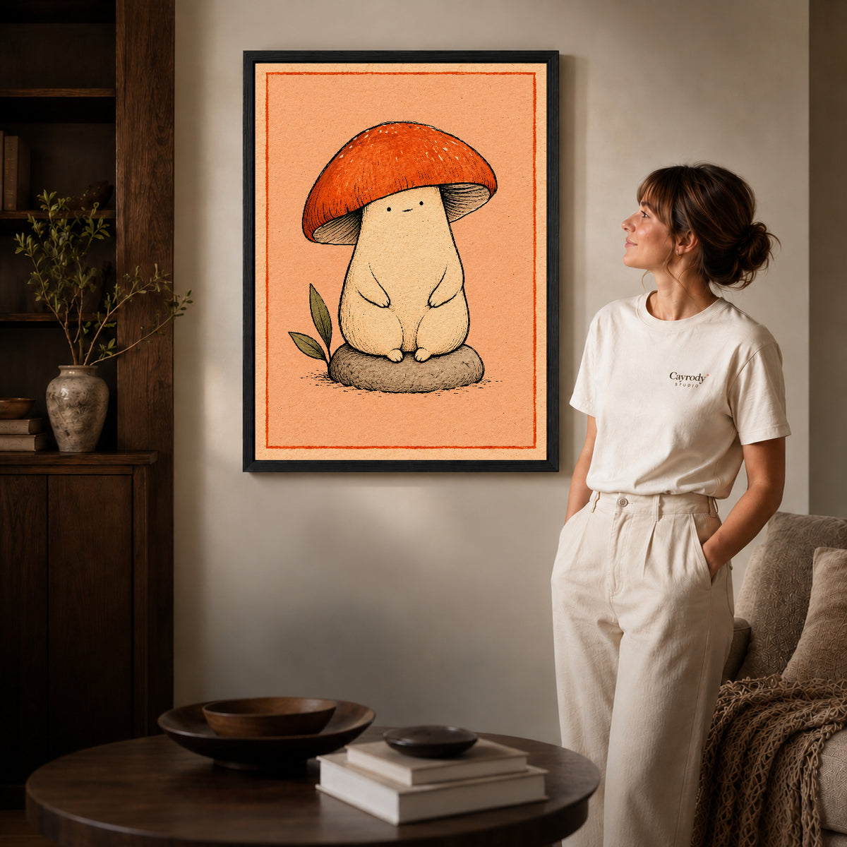cozy mushroom friend canvas art in a black frame beside a woman and round wooden table.