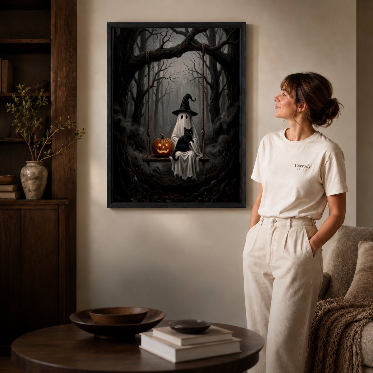 woodland ghost cat swing canvas art in a black frame beside a woman and round wooden table.