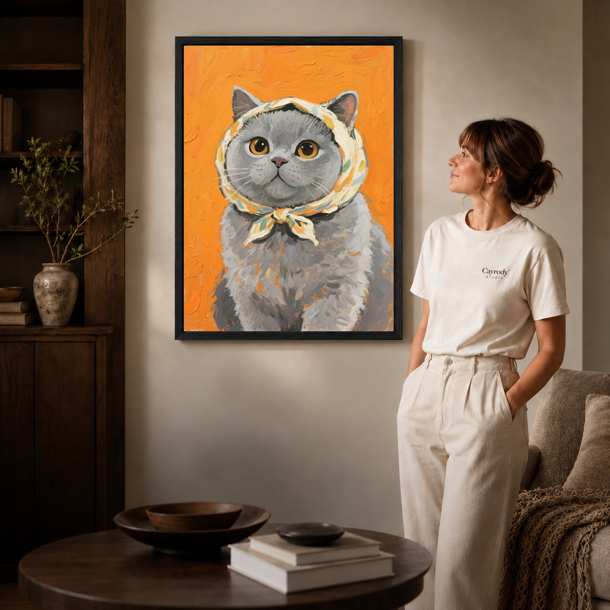 scarf wearing kitten canvas art in a black frame beside a woman and round wooden table.