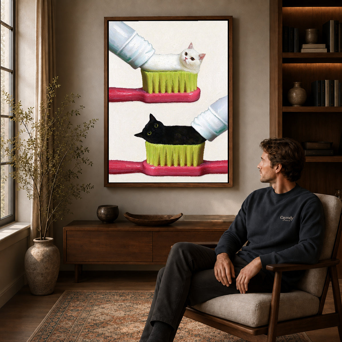 toothpaste kitten duo canvas art in a walnut frame above a console beside a seated man.