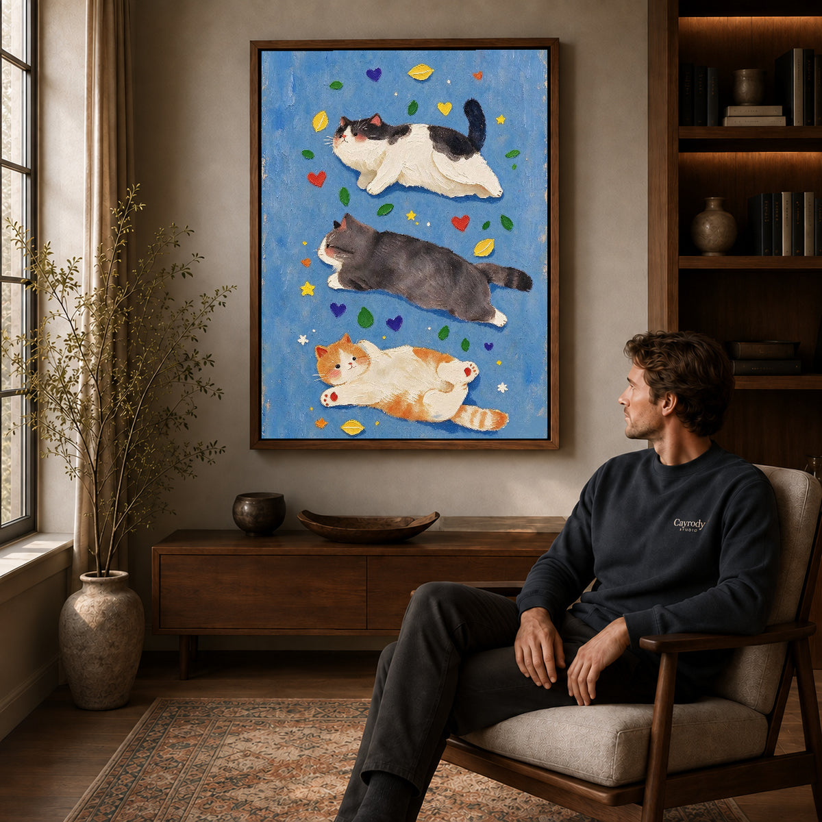 three resting cats canvas art in a walnut frame above a console beside a seated man.