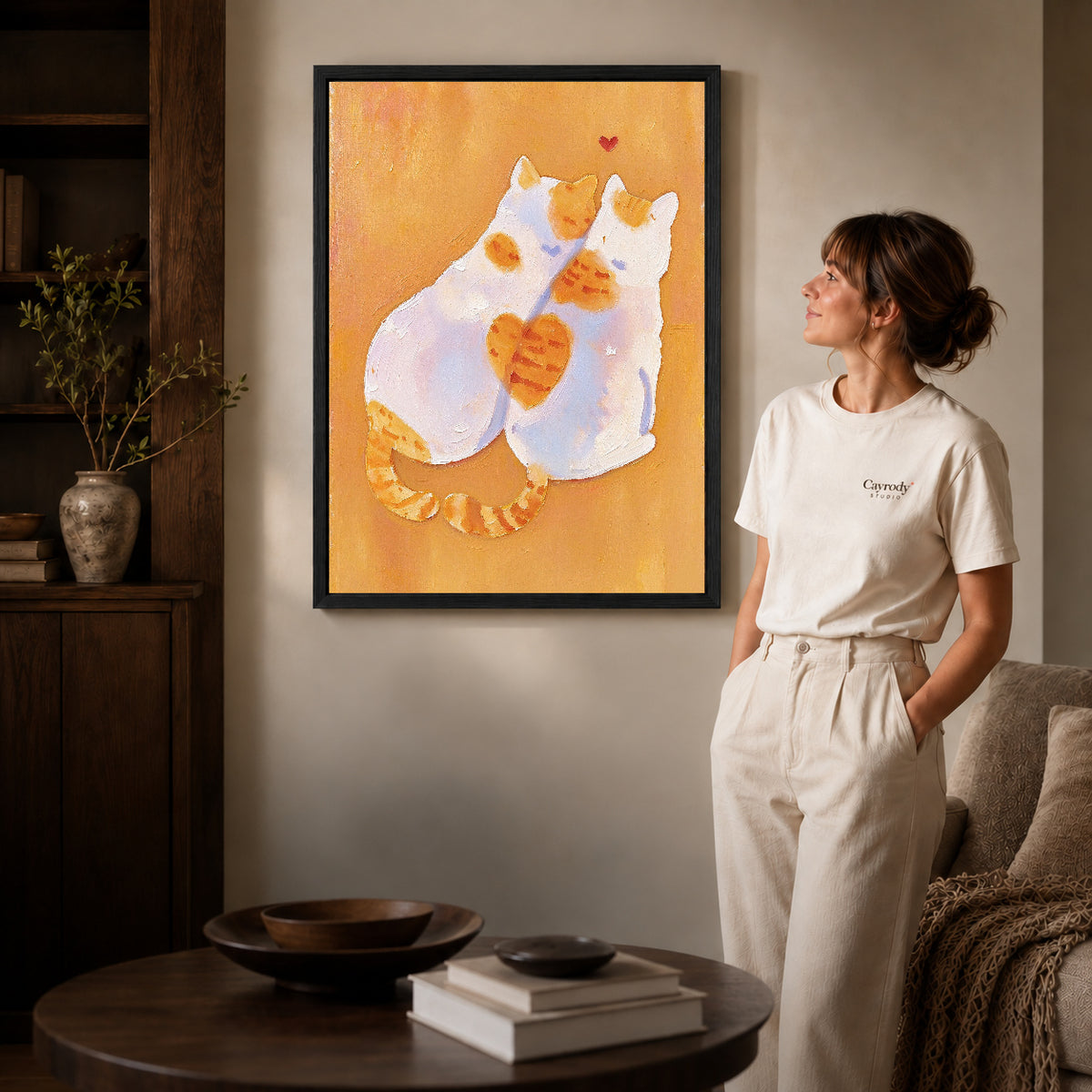 two tender cats canvas art in a black frame beside a woman and round wooden table.
