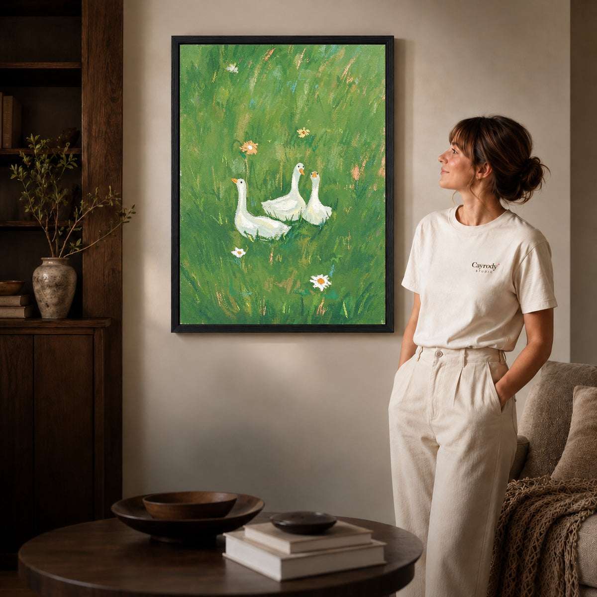 meadow goose trio canvas art in a black frame beside a woman and round wooden table.