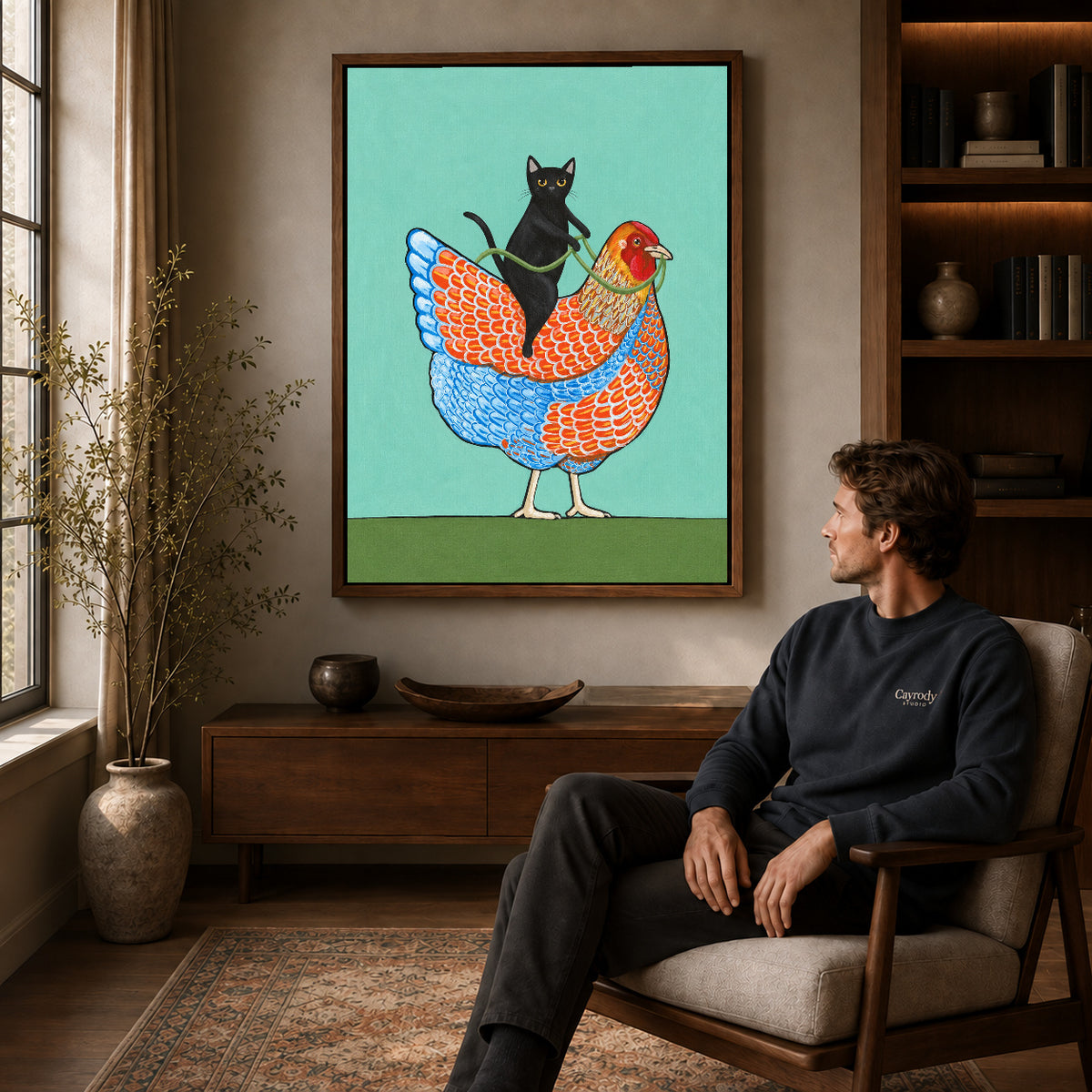 cat riding hen canvas art in a walnut frame above a console beside a seated man.