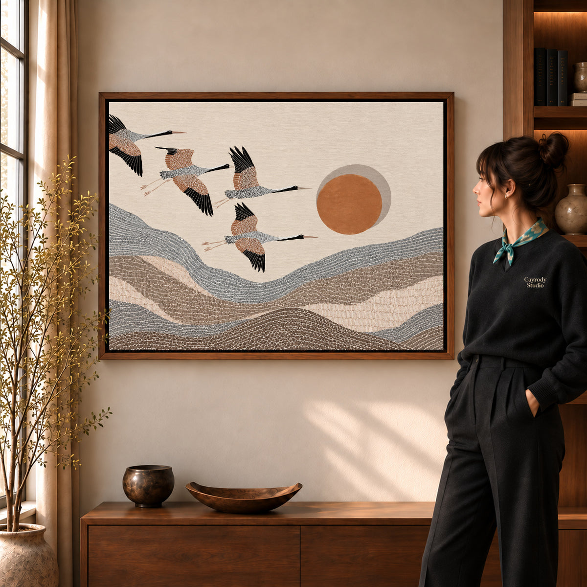 cranes mountain flight canvas art in a walnut frame beside a standing woman and wooden console.
