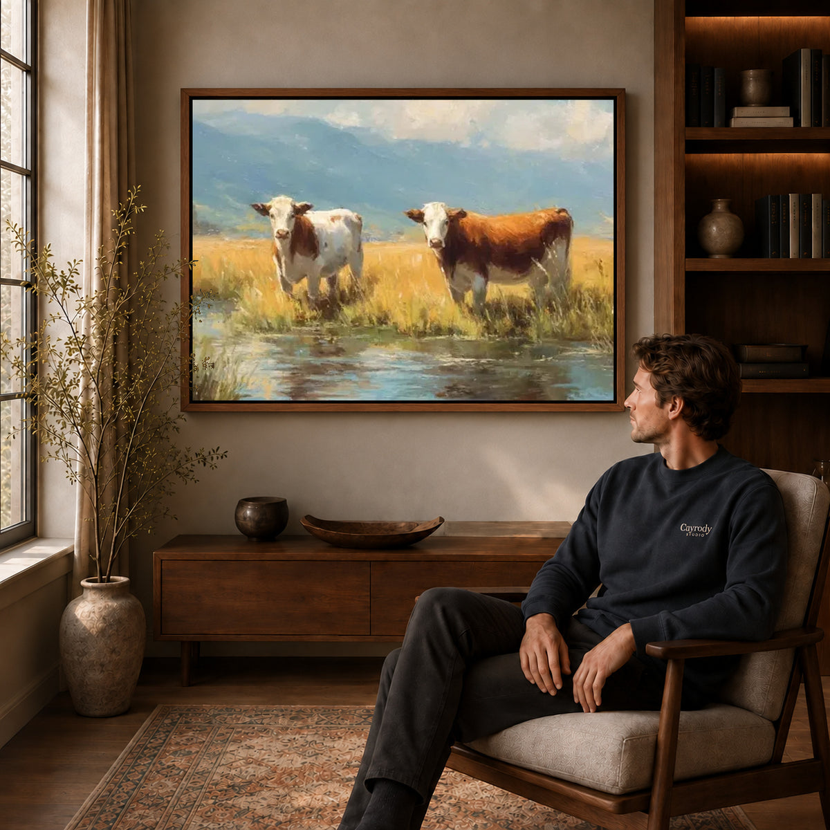 quiet meadow cows canvas art in a walnut frame above a console beside a seated man.