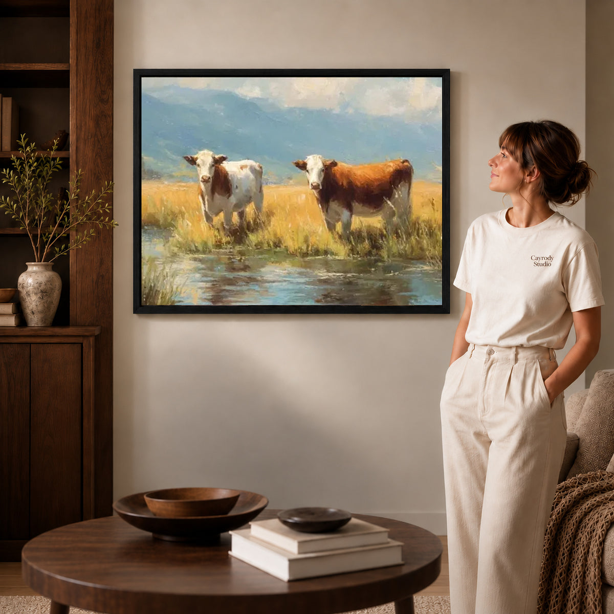 quiet meadow cows canvas art in a black frame beside a woman and round wooden table.
