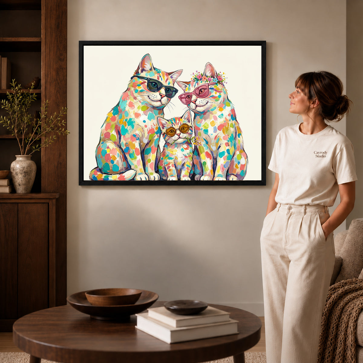 colorful cat family canvas art in a black frame beside a woman and round wooden table.