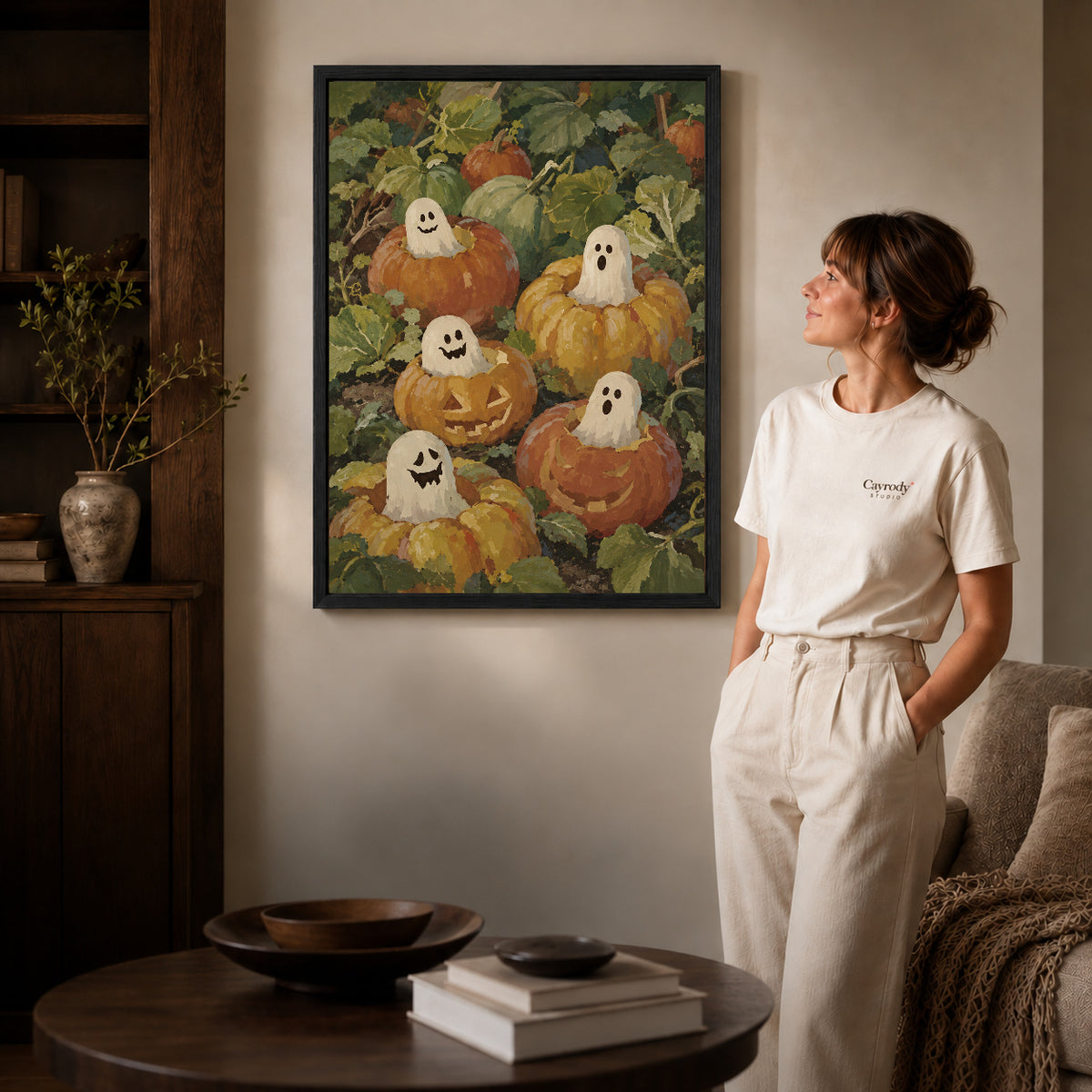 pumpkin ghost friends canvas art in a black frame beside a woman and round wooden table.