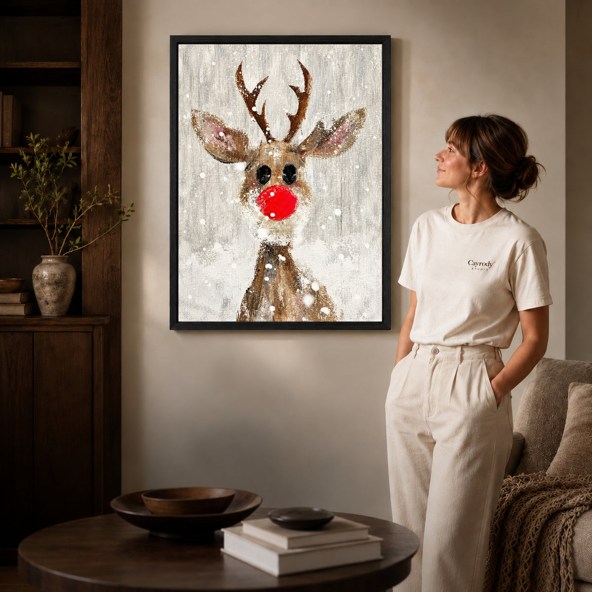 snowy red nose deer canvas art in a black frame beside a woman and round wooden table.