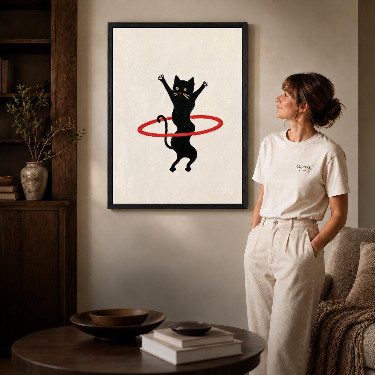 hula hoop cat canvas art in a black frame beside a woman and round wooden table.
