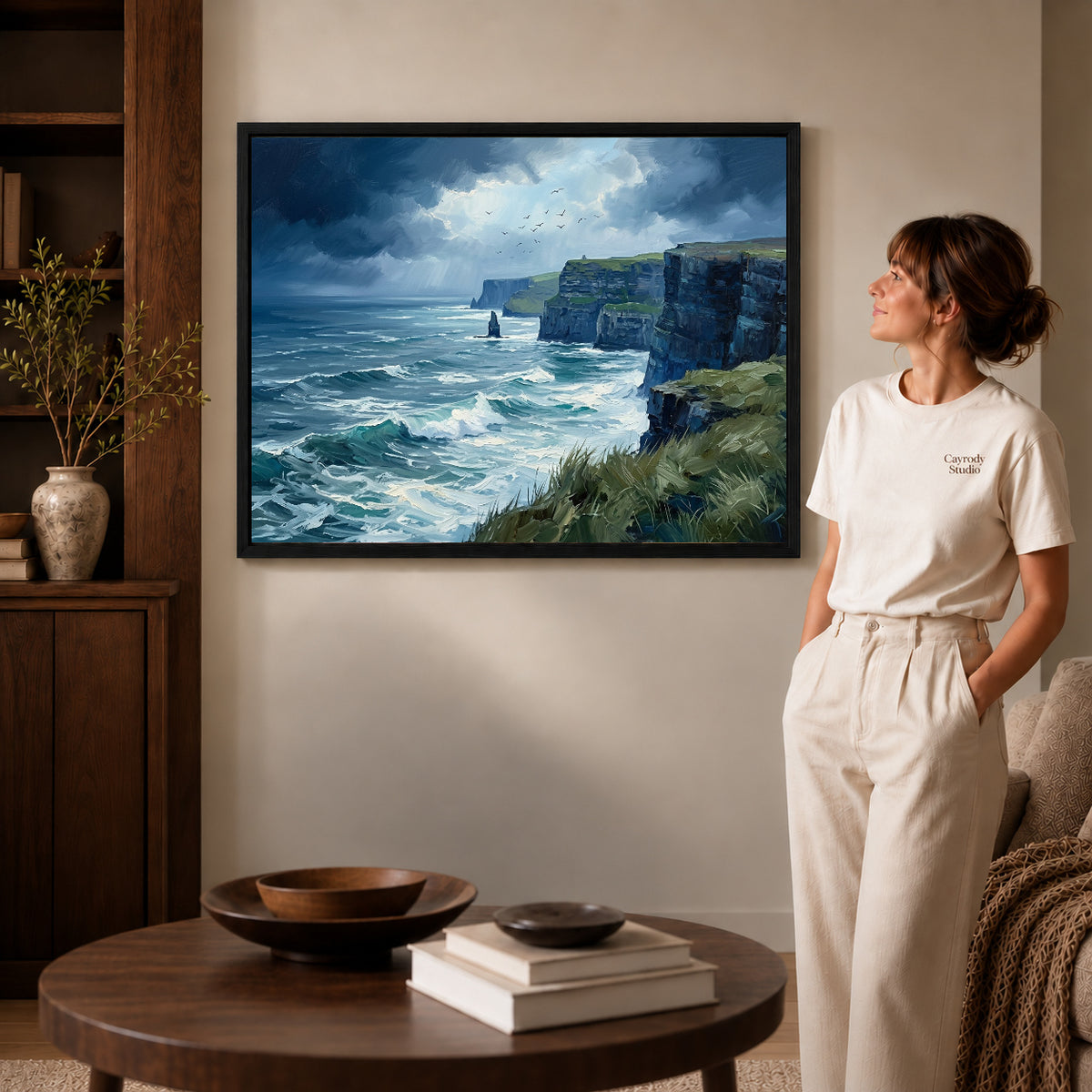 wild coastal cliffs canvas art in a black frame beside a woman and round wooden table.