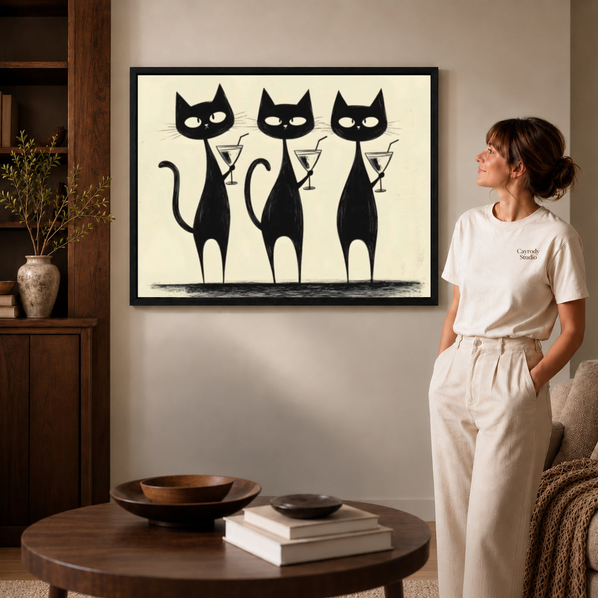 cocktail cat trio canvas art in a black frame beside a woman and round wooden table.