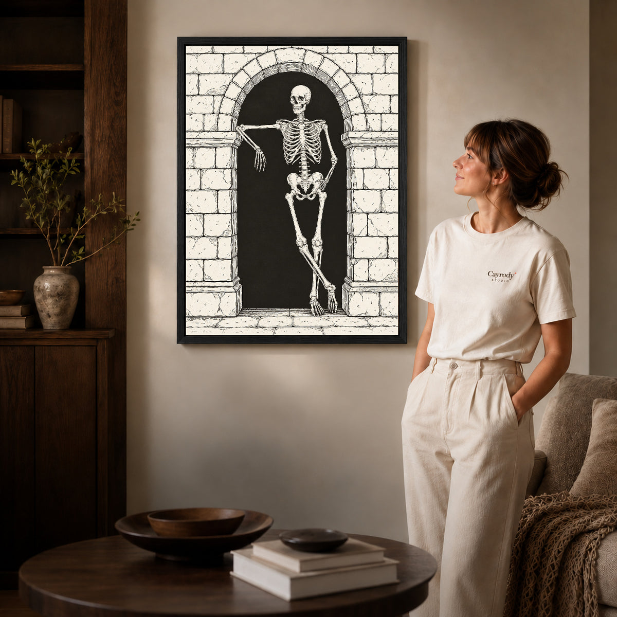 leaning skeleton guest canvas art in a black frame beside a woman and round wooden table.